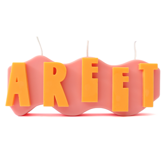 A decorative candle with a wavey body and protruding text. Measuring 17cm long and 7cm high. The body of the candle is coral pink with orange raised letters spelling AREET from left to right. It has three wicks spread across the top of the body evenly. 