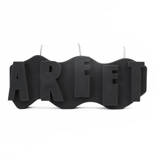 A decorative candle with a wavey body and protruding text. Measuring 17cm long and 7cm high. The  candle is charcoal black with raised letters spelling AREET from left to right. It has three wicks spread across the top of the body evenly. 
