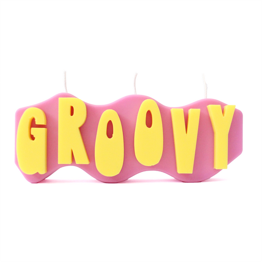A decorative candle with a wavey body and protruding text. Measuring 17cm long and 7cm high. The body of the candle is purple with bright yellow raised letters spelling GROOVY from left to right. It has three wicks spread across the top of the body evenly. 