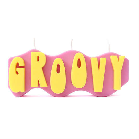 A decorative candle with a wavey body and protruding text. Measuring 17cm long and 7cm high. The body of the candle is purple with bright yellow raised letters spelling GROOVY from left to right. It has three wicks spread across the top of the body evenly.